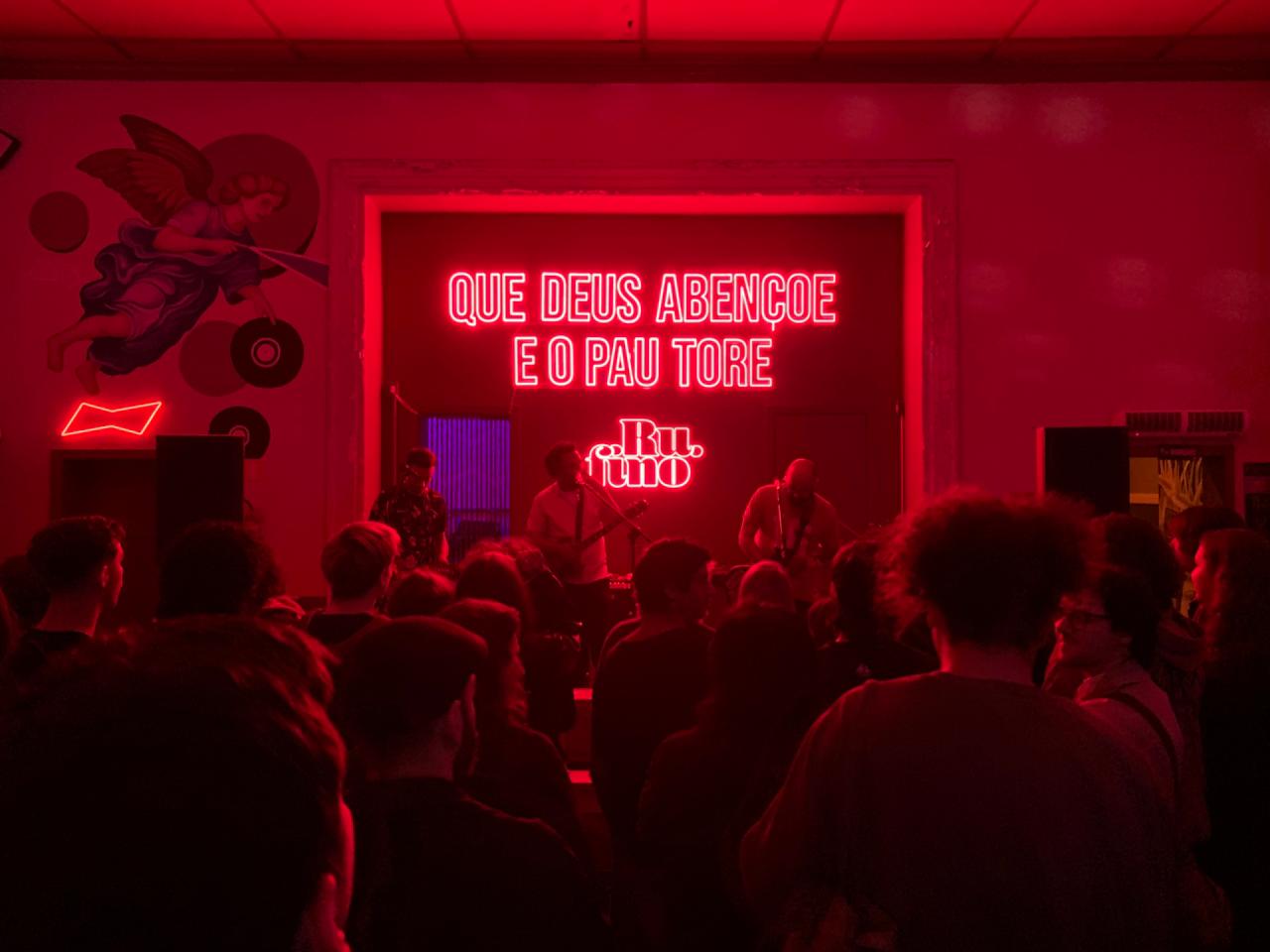 Uma multidão assiste a uma apresentação musical em um ambiente com iluminação vermelha intensa. No fundo, um letreiro de neon vermelho exibe a frase "QUE DEUS ABENÇOE E O PAU TORE" e a palavra "RUA" abaixo. À esquerda, há uma pintura de um anjo e discos de vinil na parede, enquanto à direita, equipamentos de som e decoração adicional são visíveis. A atmosfera é vibrante e cheia de energia.