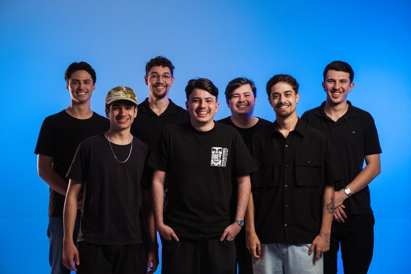 O ChatGPT disse: Um grupo de sete jovens posa sorridente diante de um fundo azul. Todos usam roupas casuais em tons escuros, transmitindo um ar descontraído e moderno. Eles estão lado a lado, alguns com as mãos nos bolsos e outros com os braços soltos, reforçando a atmosfera de união e camaradagem.