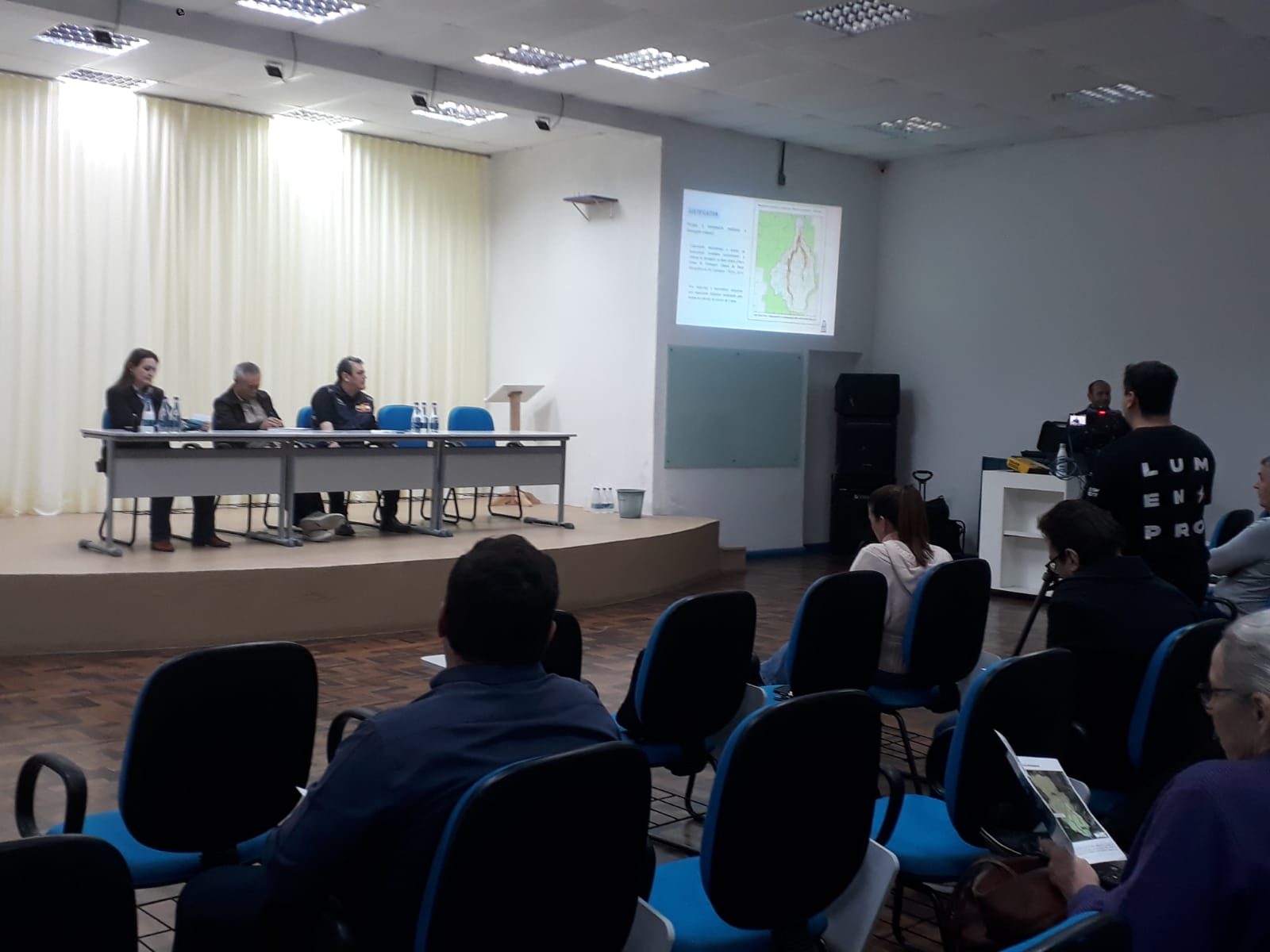 A fotografia retrata um audiencia em andamento dentro de um auditório. A composição se trata de uma audiência pública, estudos ambientais da bacia do rio Itaum-Açu, dado o conteúdo projetado no slide.