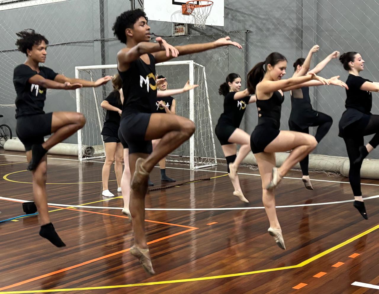 Grupo de bailarinos executa saltos sincronizados em um ginásio, elevando uma perna e mantendo os braços posicionados para a coreografia