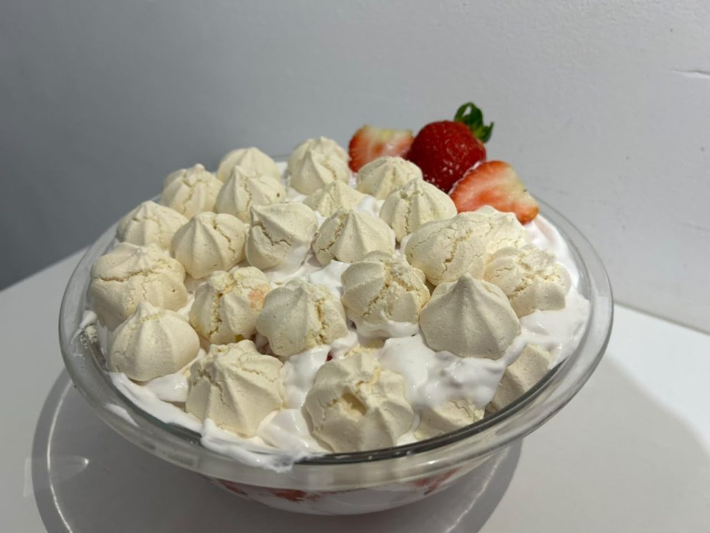 A imagem mostra uma taça de vidro transparente com Eton Mess: camadas de chantilly bem branco, pedaços de suspiro (meringue) inteiros na parte de cima, formando uma espécie de "coroa" crocante, e morangos frescos cortados ao meio aparecendo na lateral da taça. A foto está em ângulo levemente rotacionado, com fundo neutro em tons de cinza/branco e boa luz natural, destacando a textura irregular dos suspiros contra o creme.