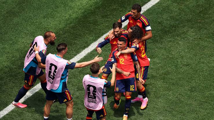 Jogadores da seleção espanhola se abraçam e comemoram em campo durante partida da Copa do Mundo 2026, com membros da comissão técnica correndo em direção ao grupo.
