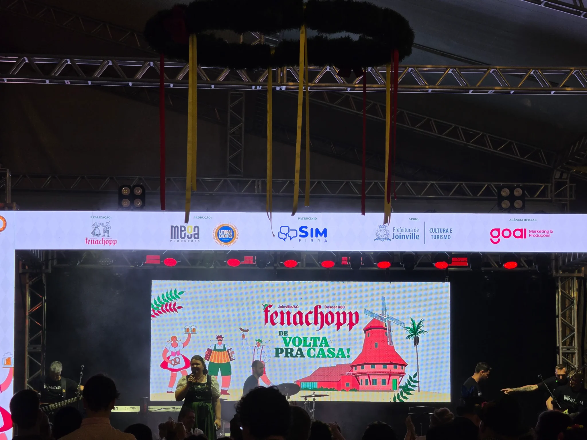 Palco principal da Fenachopp 2026, com uma mulher fazendo um anúncio em frente ao telão, com o KV do evento "Fenachopp: De Volta para Casa".