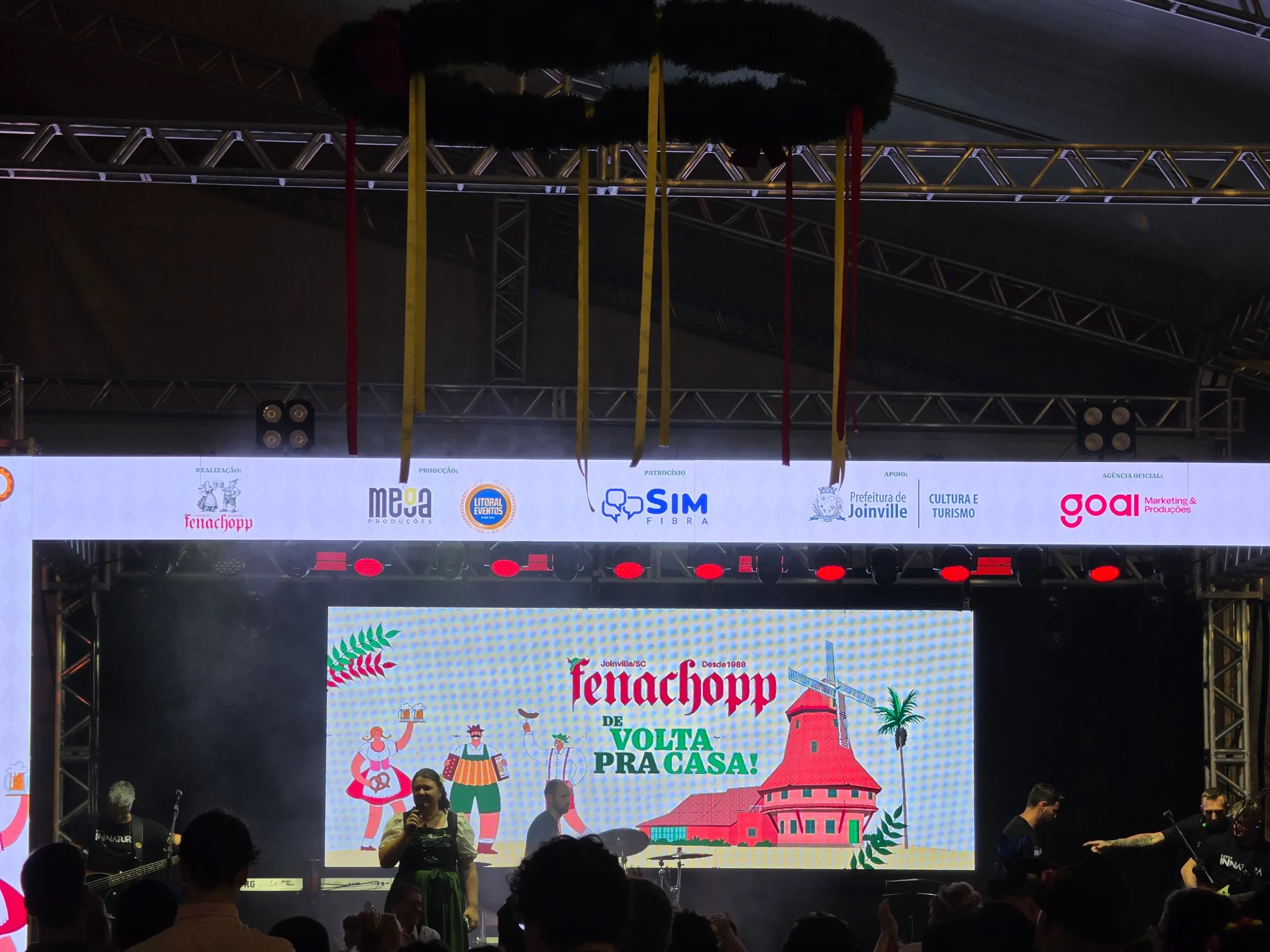 Palco principal da Fenachopp 2026, com uma mulher fazendo um anúncio em frente ao telão, com o KV do evento "Fenachopp: De Volta para Casa".