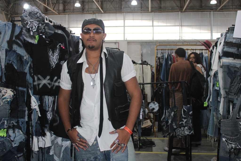 Homem negro de boné e óculos escuros, posando em frente ao seu estande de peças upcycling.