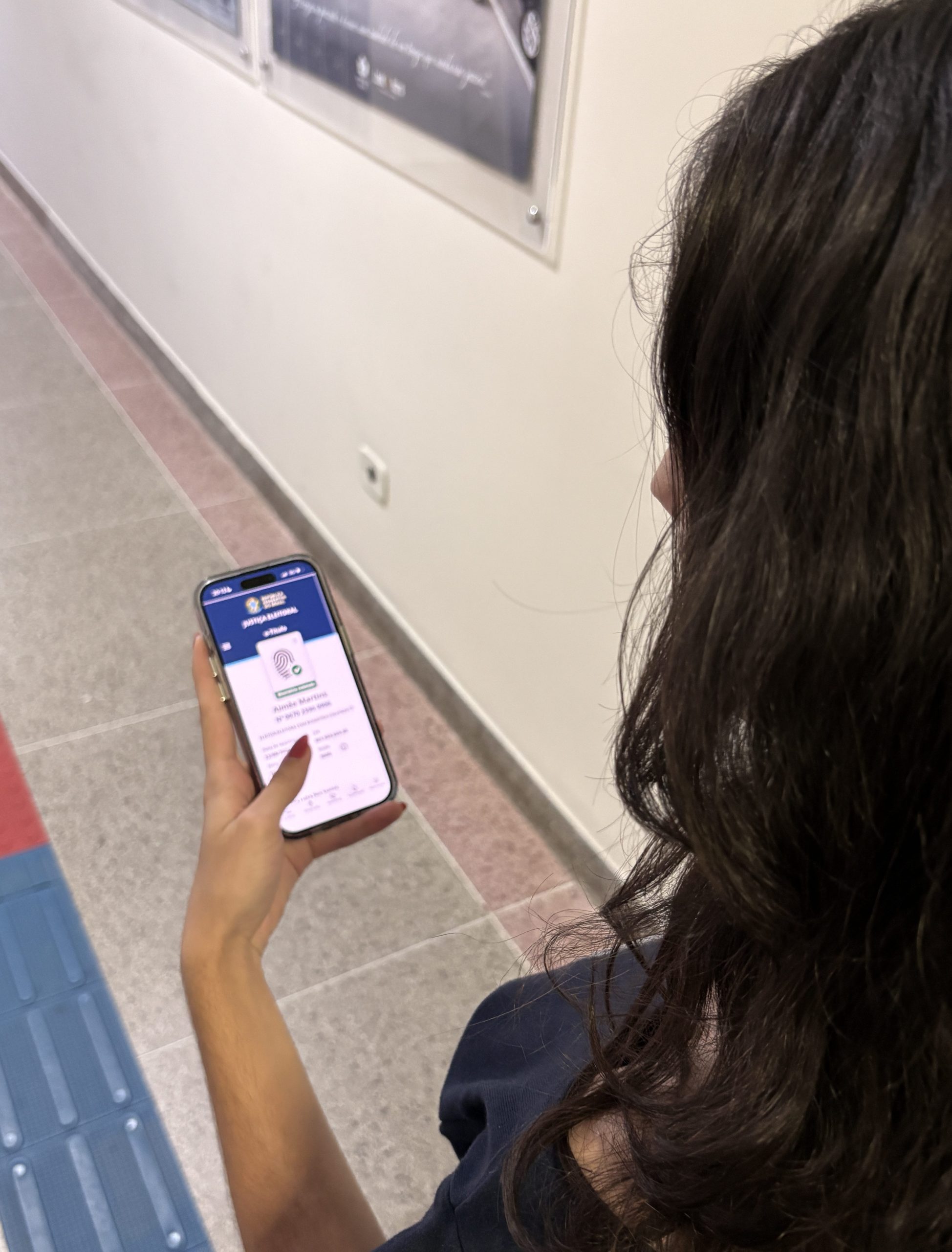 Mulher de costas, cabelo castanho, branca. Por cima de seu ombro visualizasse um celular ligado na pagina da justiça federal eleitoral do Brasil.