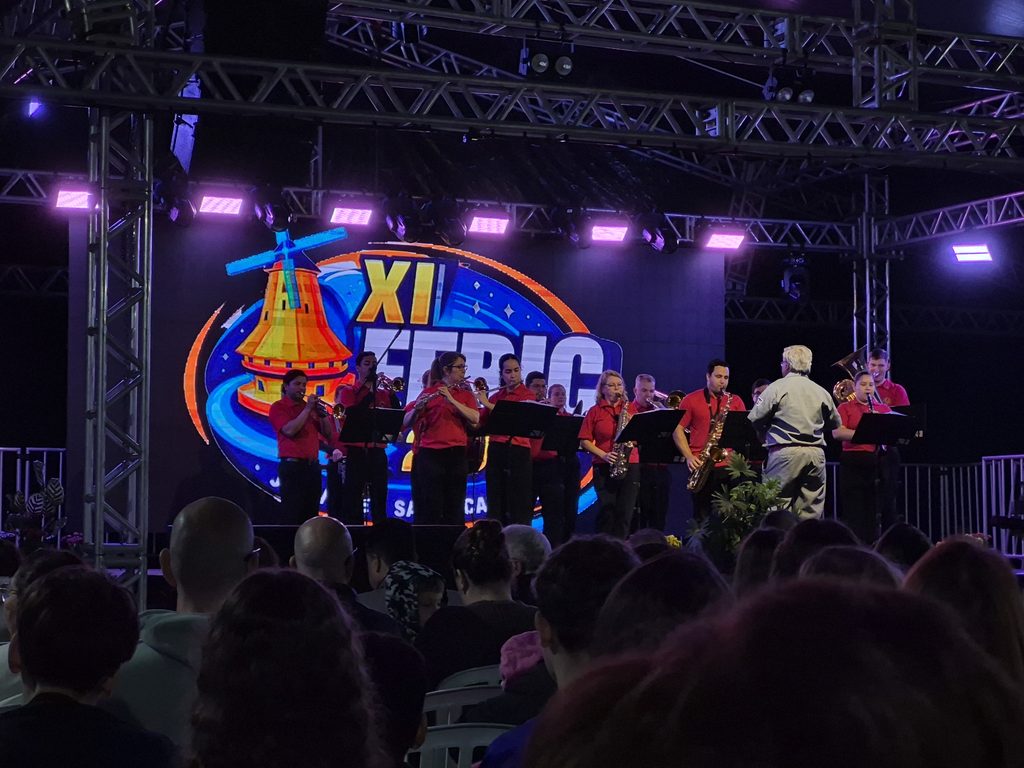 Banda se apresenta em um palco iluminado, com músicos tocando instrumentos de sopro. Ao fundo, um painel colorido exibe “XI FEBIC”, enquanto o público acompanha a apresentação.