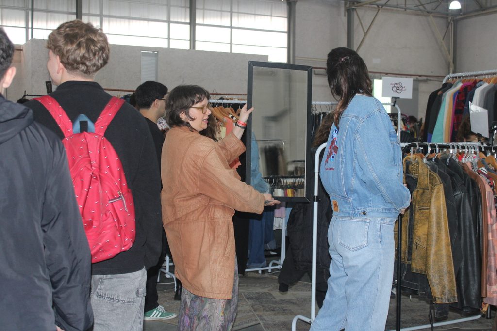 Mulher branca de cabelo castanho experimentando uma jaqueta jeans com desenho nas costas, olhando para o espelho que uma mulher segura.