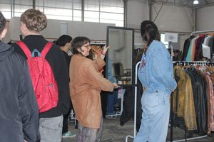Mulher prova jaqueta jeans enquanto se olha no espelho, enquanto uma outra mulher o segura. Pessoas circulam ao fundo.
