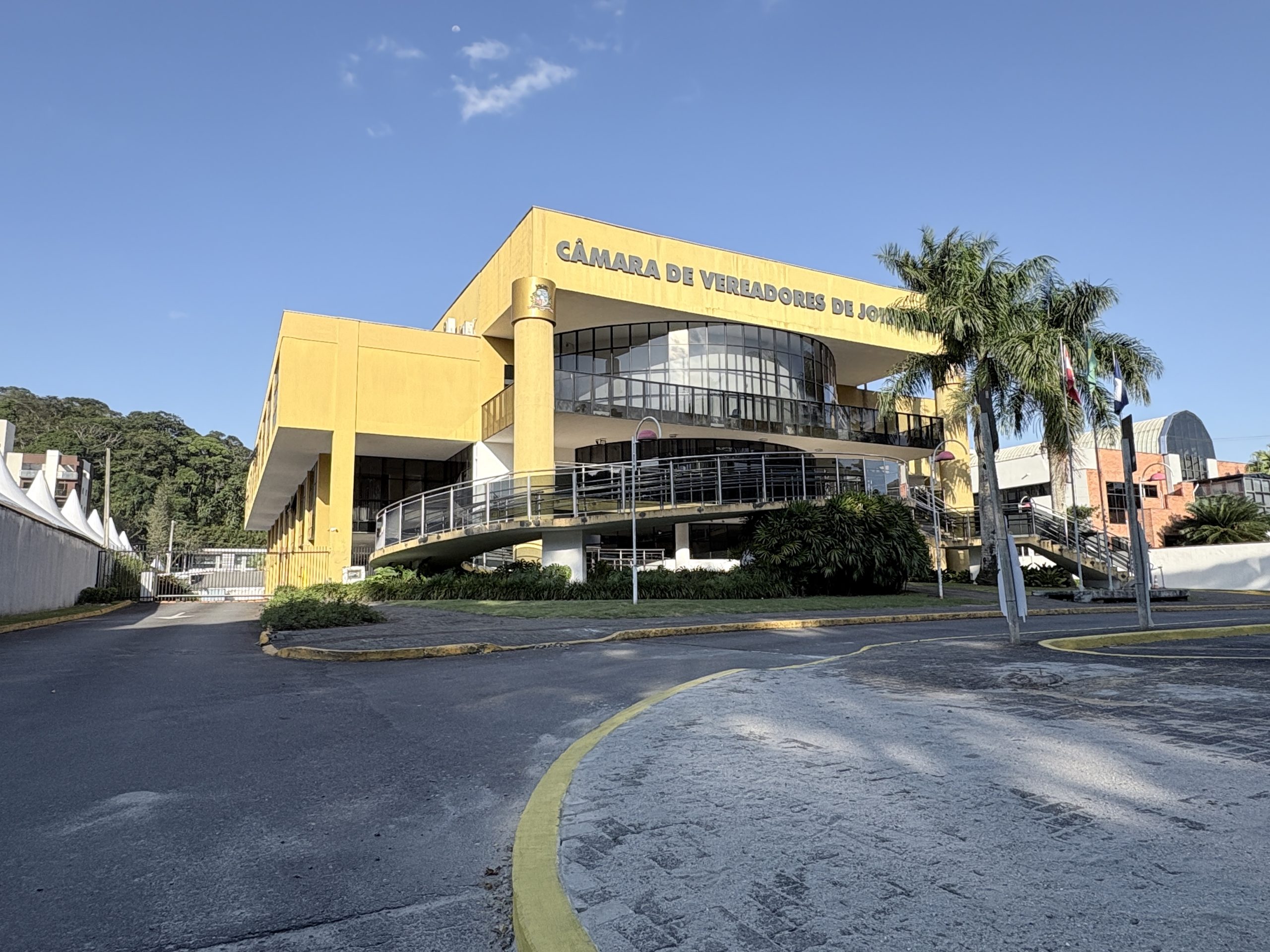 Faixada da câmara dos vereadores de Joinville. O prédio é amarelo, com janelas de vidro na frente e escadaria acessivel compondo sua fachada. Tem um caminho asfaltado de estacionamento a esquerda e calçada na frente. onde também tem um jardim de grama na frente para decorar. Tem duas palmeiras na frente e as bandeiras hasteadas do Brasil, de Santa Catarina e de Joinville.