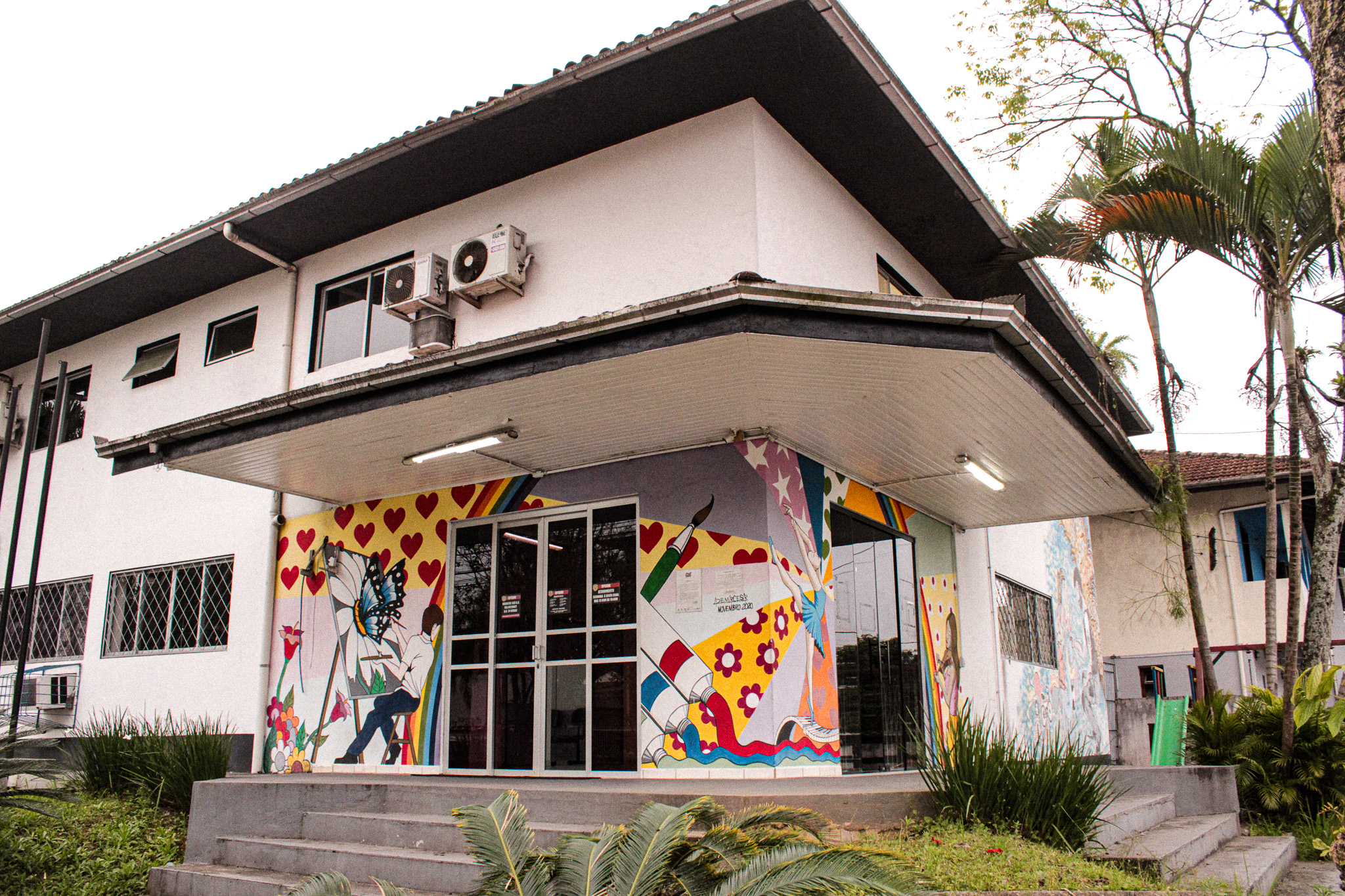 Fachada de prédio branco de dois andares da Delegacia da Mulher em Joinville, com entrada decorada por grafite colorido com corações e pincéis.