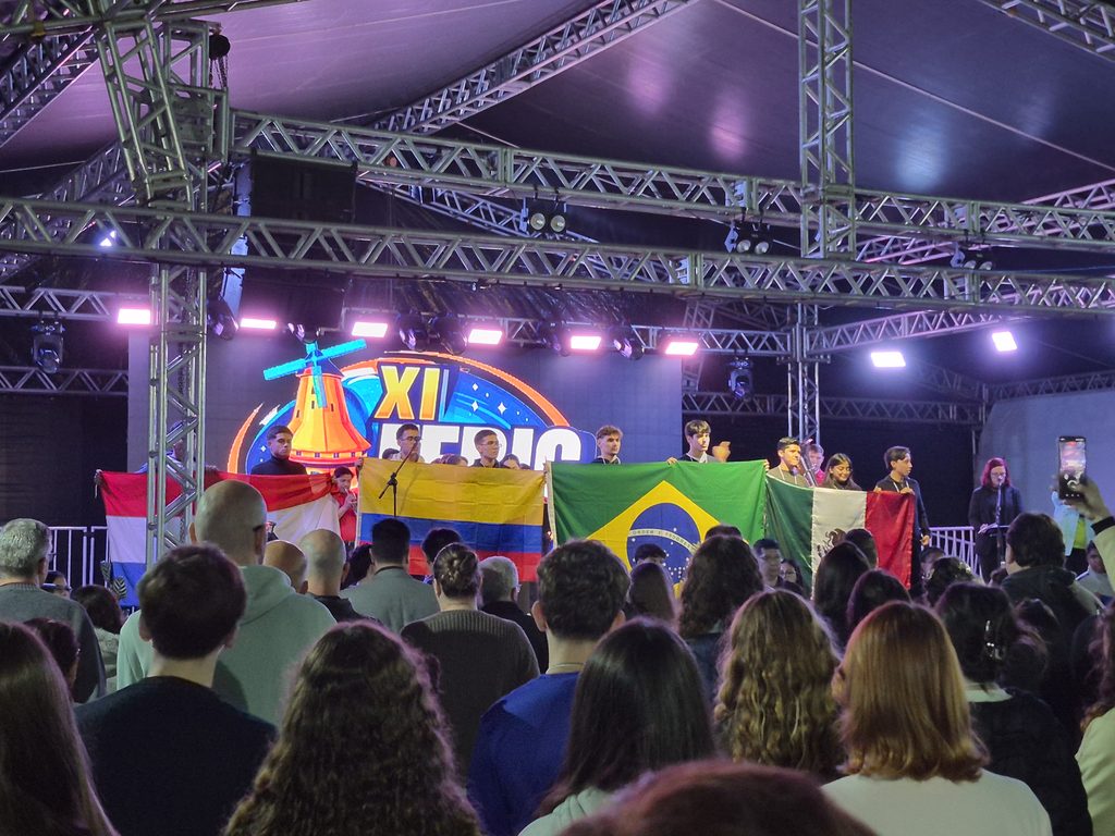 No palco iluminado, com um um painel colorido exibindo “XI FEBIC”, jovens estudantes seguram as bandeiras do México, Paraguai, Colômbia e Brasil diante de uma plateia.