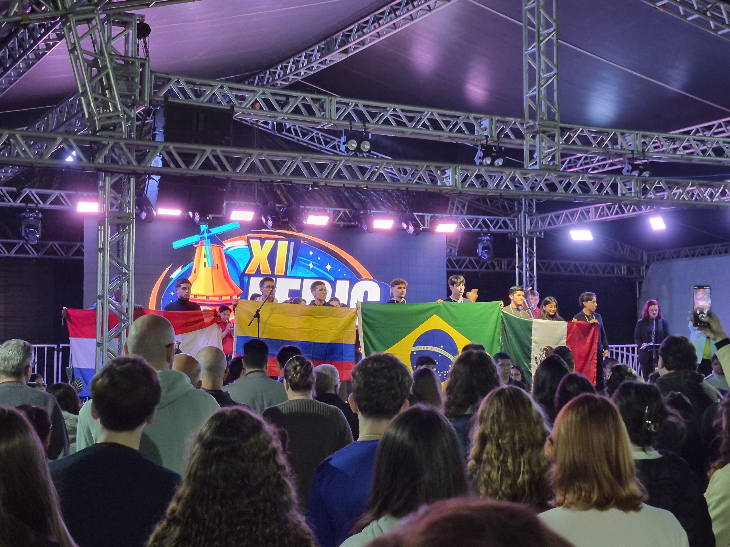 No palco iluminado, com um um painel colorido exibindo “XI FEBIC”, jovens estudantes seguram as bandeiras do México, Paraguai, Colômbia e Brasil diante de uma plateia.