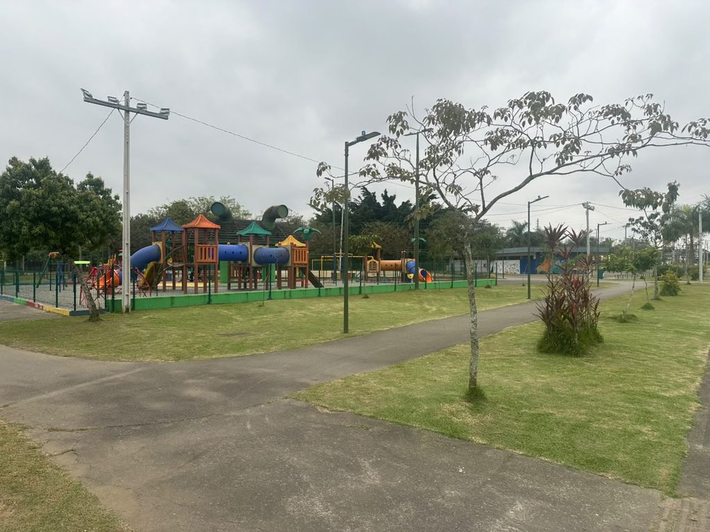 Fotografia diurna do Parque da Cidade sob um céu nublado. Em primeiro plano, há caminhos asfaltados cercados por gramado verde e pequenas árvores. Ao fundo, protegido por uma grade verde, há um grande parquinho infantil colorido com diversos escorregadores e estruturas para escalada.
