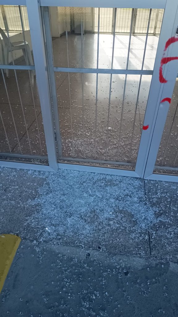 Fotografia mostrando a parte inferior de uma porta de vidro e metal. O chão de concreto em frente à porta está completamente repleto de pequenos cacos de vidro temperado estilhaçado. Na lateral da esquadria da porta, há marcas de pichação em tinta spray vermelha.