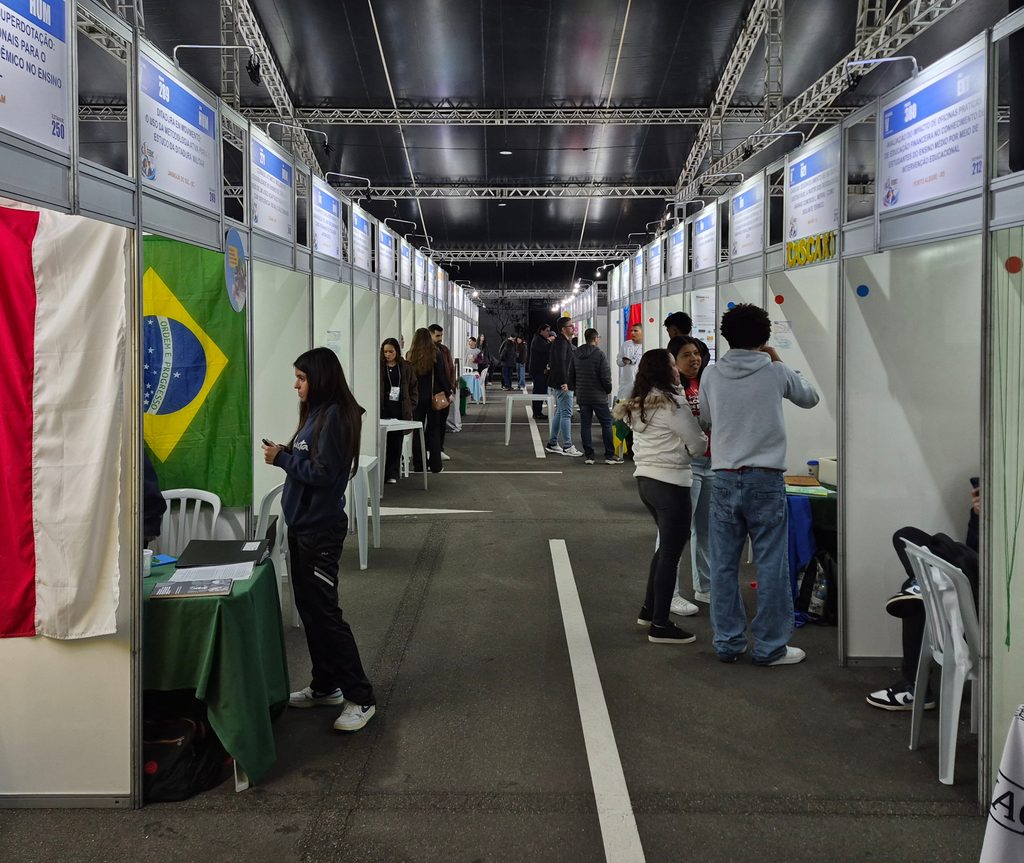 Corredor coberto com fileiras de estandes brancos dos dois lados. Estudantes e visitantes conversam em pé entre as bancadas.
