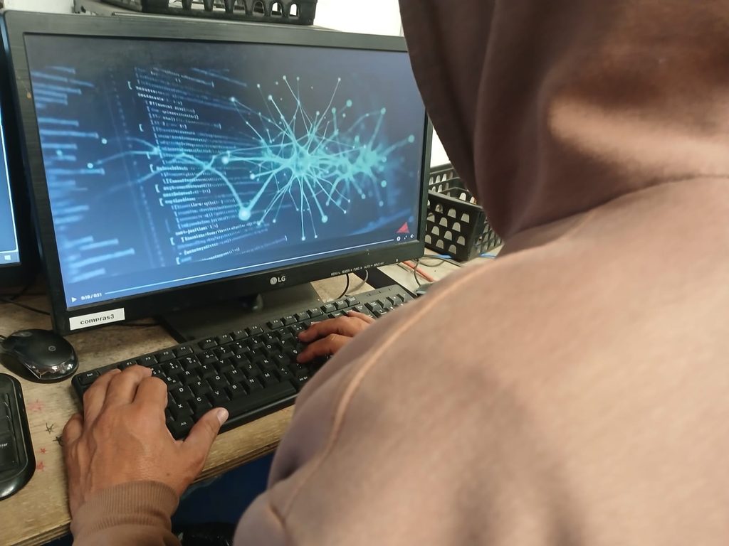Pessoa de costas usando moletom com capuz em um quarto escuro, digitando em um computador que exibe telas com dados e uma janela com o título