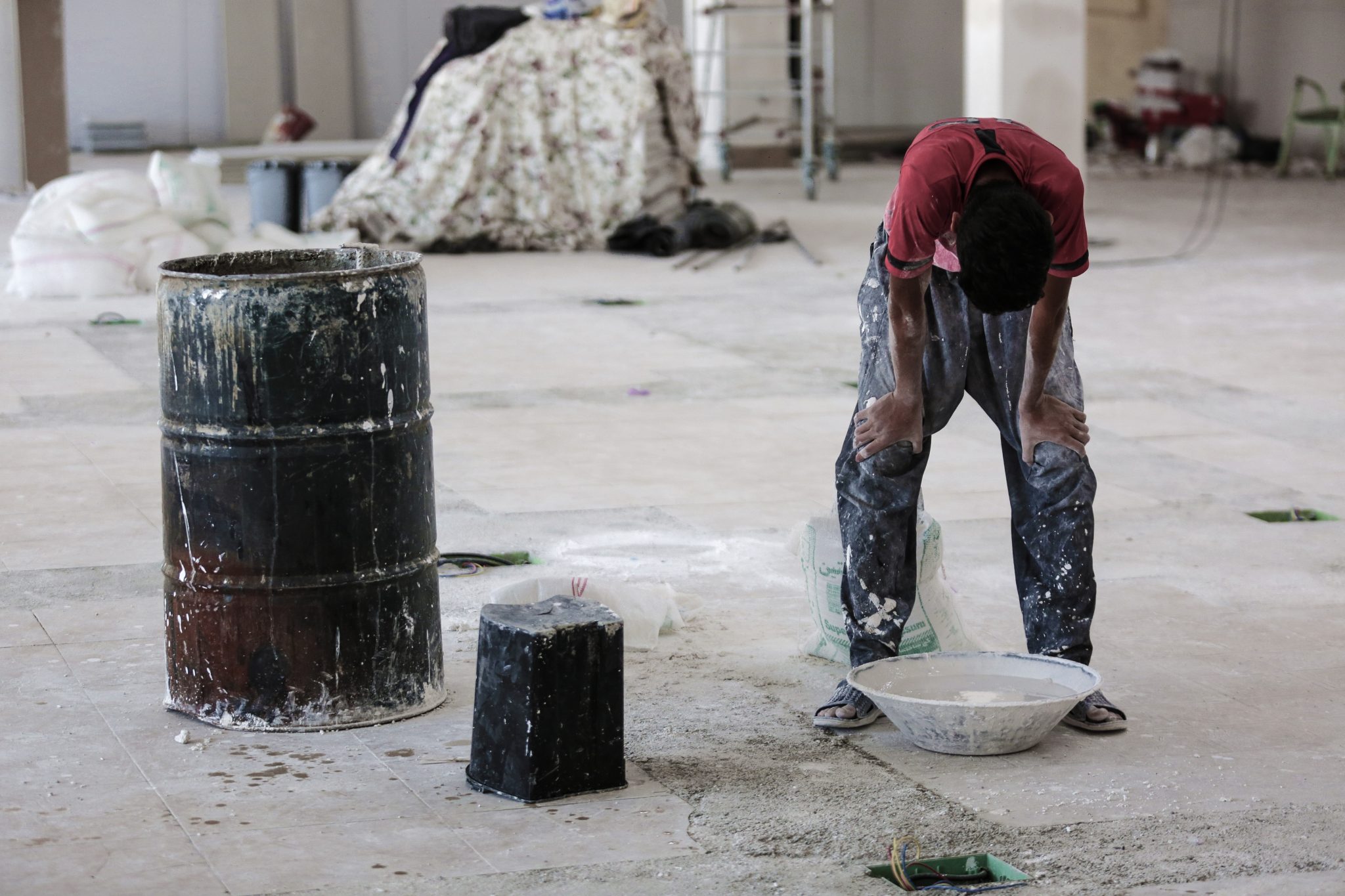 Fotografia mostra um trabalhador em um ambiente de construção, curvado enquanto apoia as mãos sobre os joelhos. Ele veste camiseta vermelha, calça manchada de tinta e chinelos. Ao redor, há um tambor, baldes, sacos de material e ferramentas espalhadas pelo chão.