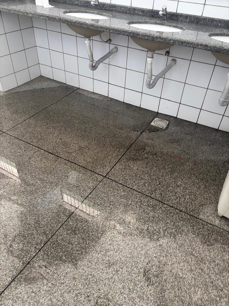 Fotografia do interior de um banheiro. O chão de granito escuro está coberto por uma grande poça d'água, indicando alagamento. Ao fundo, vê-se a parte inferior de uma bancada com três pias e a parede revestida com azulejos brancos.