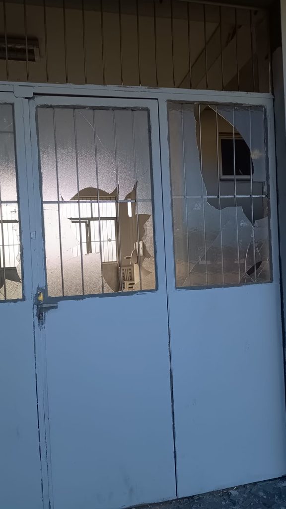 Fotografia de uma porta ou janela de metal pintada de azul claro com painéis de vidro. Vários desses vidros estão severamente quebrados, com grandes buracos e estilhaços pendurados. Atrás dos vidros, há grades de segurança de metal.
