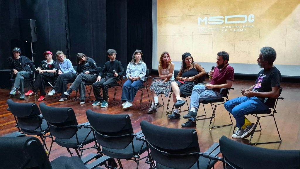 Em um auditório, dez pessoas estão sentadas em cadeiras dispostas em semicírculo sobre um palco de madeira. Duas delas seguram microfones, enquanto as demais acompanham a conversa. Ao fundo, uma tela exibe a identidade visual da IX Mostra Sesc de Cinema. Na frente do palco, há fileiras de cadeiras vazias voltadas para os participantes.
