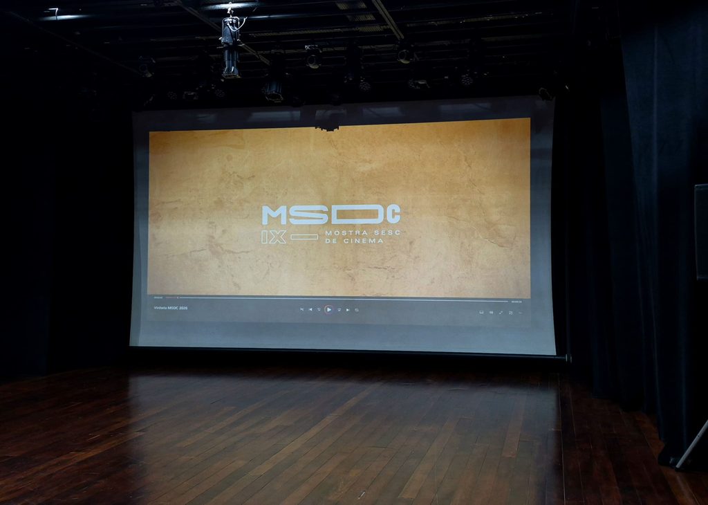 Palco vazio com piso de madeira escura e cortinas pretas nas laterais. Ao centro, uma grande tela de projeção exibe um fundo amarelo-ocre com o logotipo da IX Mostra Sesc de Cinema. O ambiente está escuro, com equipamentos de iluminação visíveis no teto.