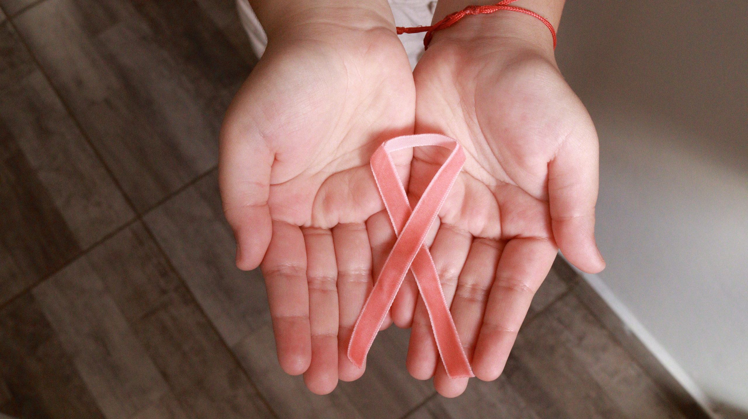 Duas mãos em concha seguram um laço de fita rosa. A pessoa usa uma pulseira vermelha no pulso direito.