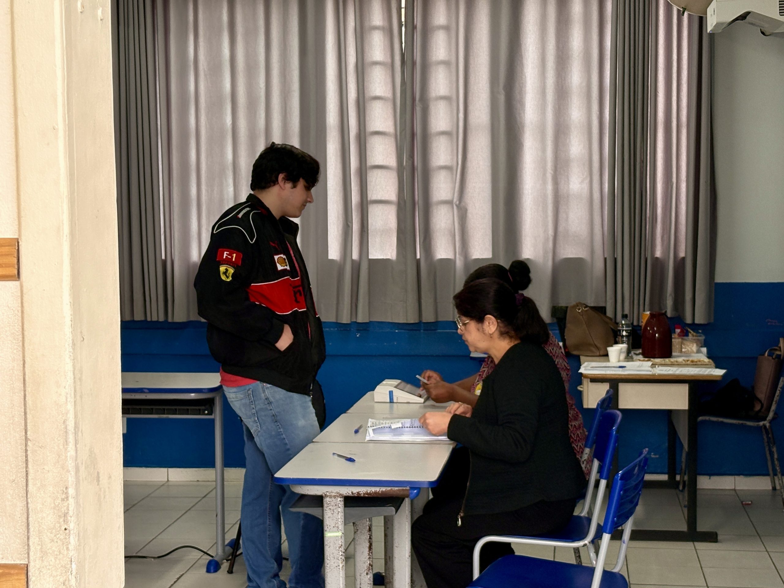 Rapaz de 22 anos de jaqueta preta da ferrari está parado em frente a duas mesárias em sua seção de votação.