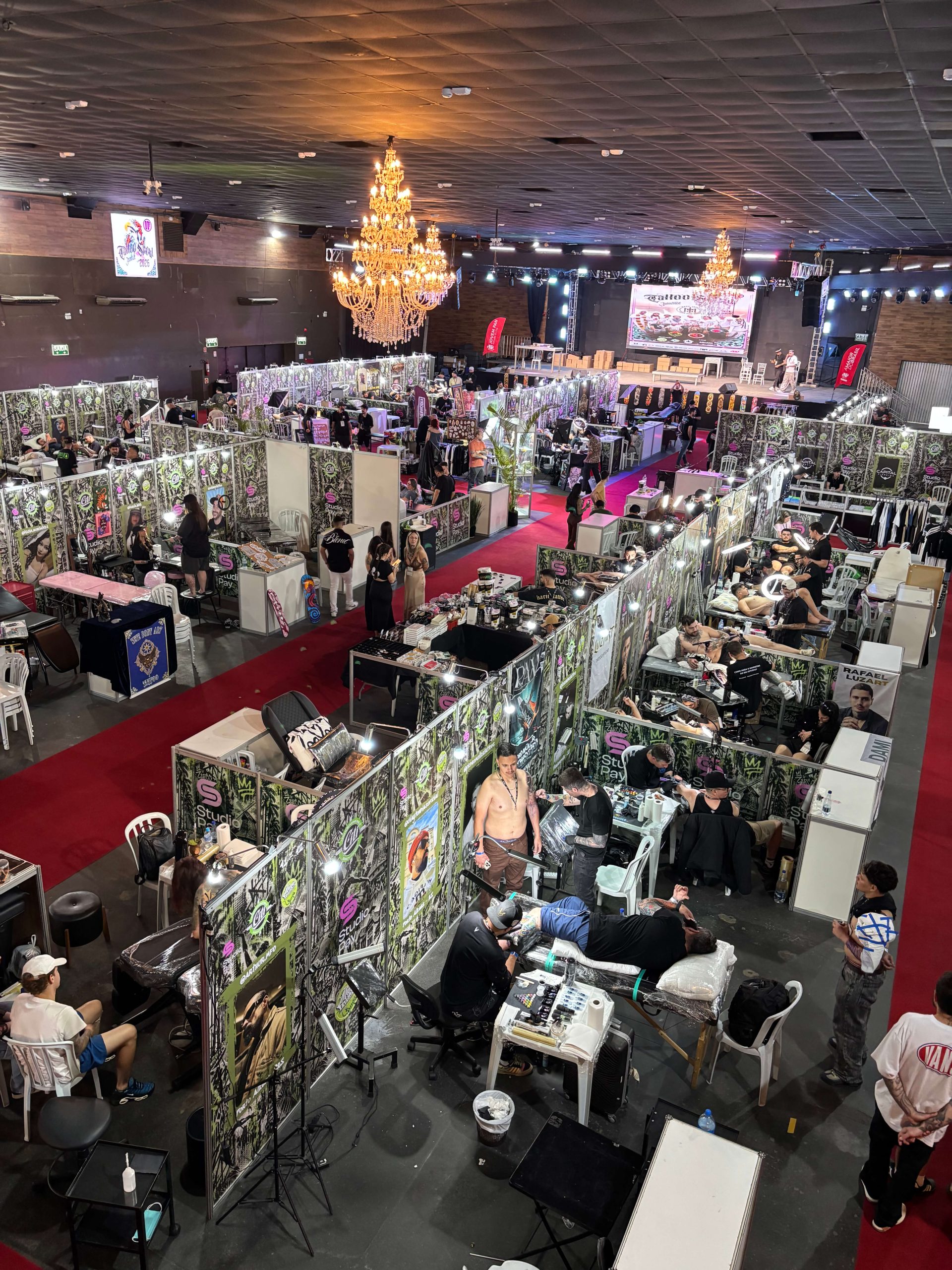 Vista panorâmica do Tattoo Show. Estandes dos tatuadores, profissionais realizando tatuagens e visitantes circulando pelo espaço do evento.