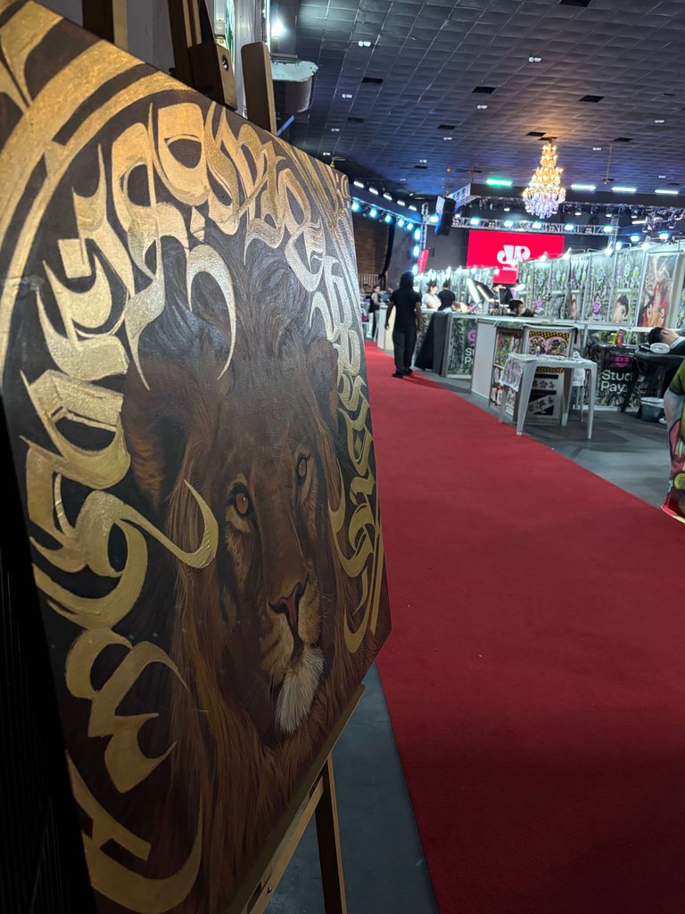 Quadro em exposição, posicionado em um cavalete sobre um tapete vermelho, retratando o rosto detalhado de um leão em tons de marrom emoldurado por complexas caligrafias e ornamentos em tinta dourada.