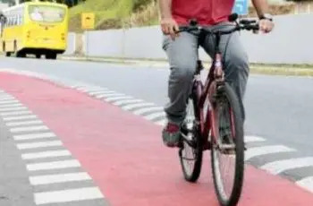 Ciclista pedalando em uma ciclofaixa vermelha, ao lado de uma avenida onde passa um ônibus amarelo.