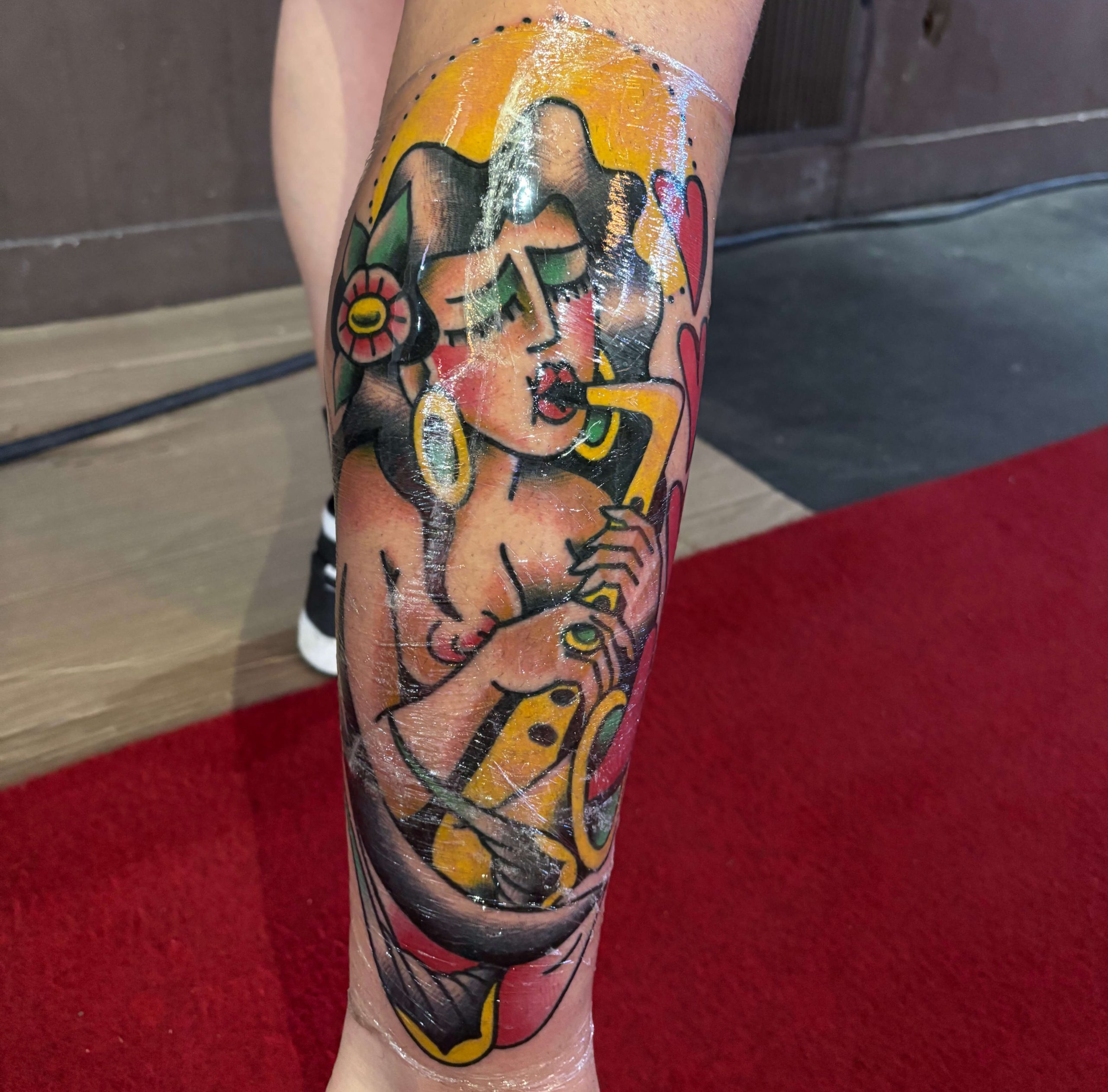Tatuagem na perna de Arno Nemitz, estilo Old School. Retrata uma mulher estilo pin-up de cabelos escuros tocando saxofone, com uma flor, argolas amarelas e corações vermelhos