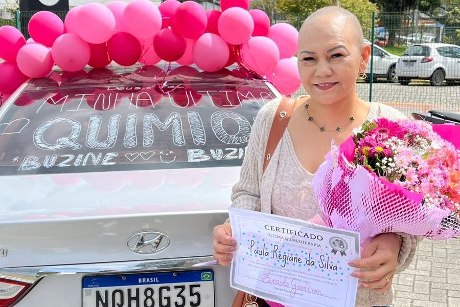 Mulher sorridente, com a cabeça raspada, segura um buquê de flores coloridas e um certificado de conclusão da última sessão de quimioterapia, em frente a um carro branco decorado com balões rosas e mensagens de carinho. A foto registra um momento de celebração e superação.