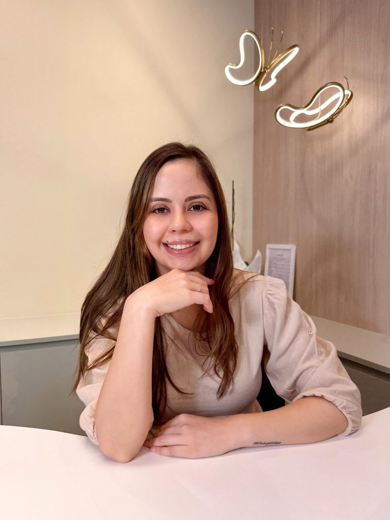 Fotografia de perfil da fonoaudióloga Ana Nascimento, da Clínica Bem te vi em Jaraguá do Sul. Ela é uma mulher jovem de cabelos castanhos compridos, está a sorrir e apoia o queixo na mão sobre uma bancada. Veste uma blusa bege de mangas bufantes e tem uma pequena tatuagem no antebraço. Ao fundo, há um painel de madeira com uma luminária decorativa em formato de borboleta.