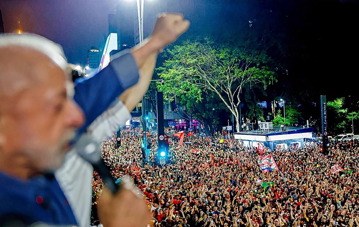 Lula aparece desfocado do lado esquerdo da foto, com a mão levantada comemorando. Na sua frente tem centenas de pessoas, com bandeiras e vestidos de vermelho