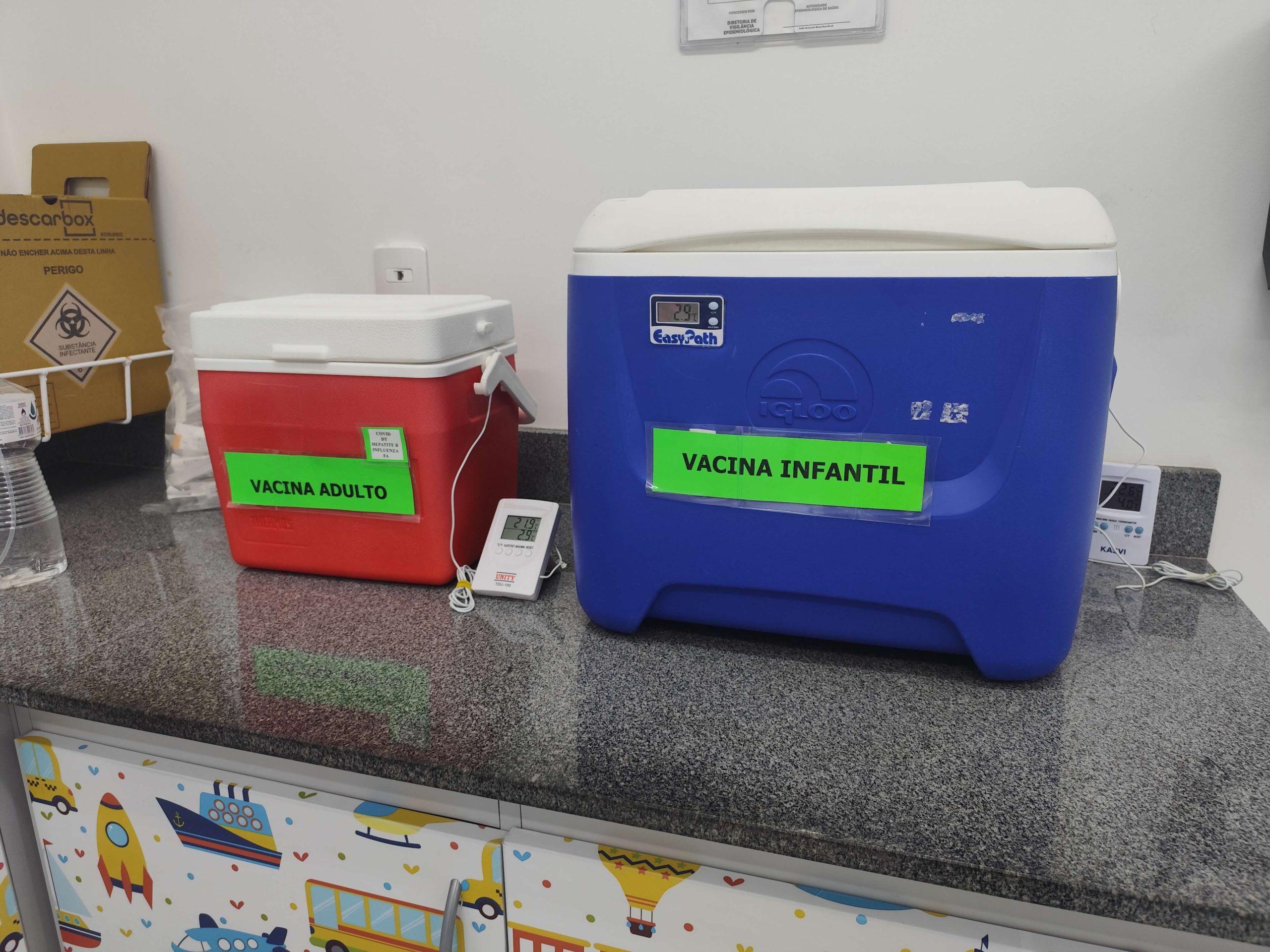 Caixas térmicas identificadas como “Vacina Adulto”, na cor vermelha, e “Vacina Infantil”, na cor azul, estão sobre uma bancada em uma unidade de saúde. Ao lado, há termômetros digitais para monitorar a temperatura dos equipamentos.